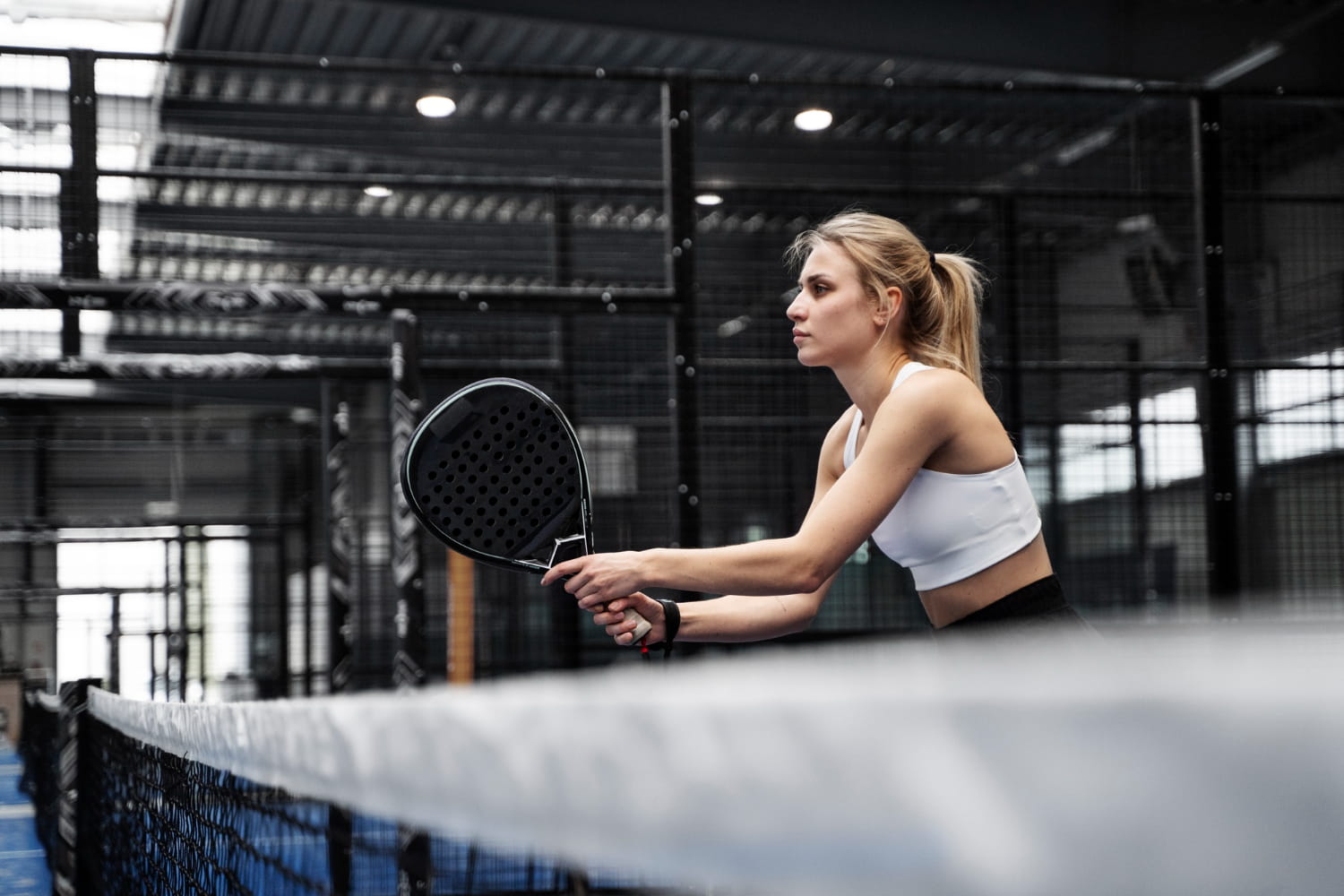 Woman holding a padel racket at an indoor court net, ideal gear available at padel shop Cyprus